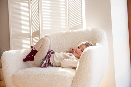 An upset little girl does not want to be photographed, turns away from the camera, covers the lens with her hand. Harmful, naughty child sits in white soft chair against backdrop of Christmas tree.の写真素材