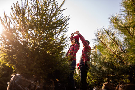 Funny couple in love in checkered red shirts knitted hats dances, spins among green Christmas tree...