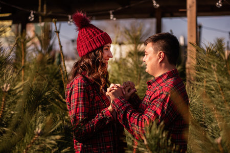 Cheerful couple in love in checkered red shirts, knitted hats hold hands among green Christmas tree market. Young man looks lovingly at beautiful woman without letting go of hands. Holiday atmosphereの写真素材