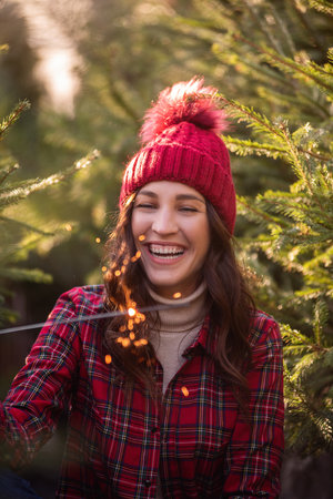 Close-up portrait of young woman with bright sparkler in hand in red plaid shirt, knitted hat among green pine trees. Curly-haired girl laughs, rejoices at New Year at Christmas tree market. Holidayの写真素材