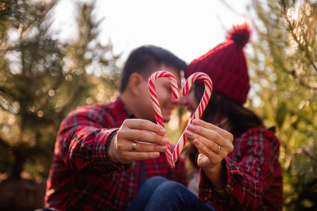 Red white candy cane is held in outstretched hands of loving couple in checkered shirts, knitted hats near green market of Christmas trees. Happy man and woman kiss, laugh. Copy spaceの写真素材