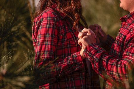Close-up of hands, loving couple in checkered red shirts holding hands tightly among green Christmas tree market. Holiday atmosphere. Concept of tactility, cohesion. Romantic winter date in the cityの写真素材