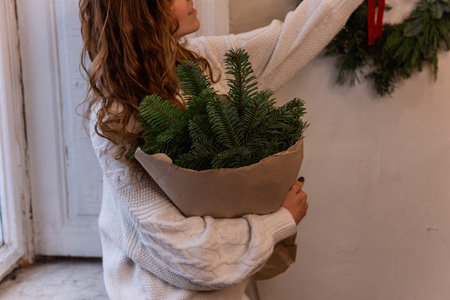 Faceless portrait of young woman in white knitted sweater, sitting on windowsill, holding in hands Christmas bouquet in craft paper near window. Eco winter decoration, wreath made from spruce branchesの写真素材