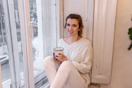 Close-up portraits of young stylish woman sitting on windowsill, drinking cocoa from cup, looking...