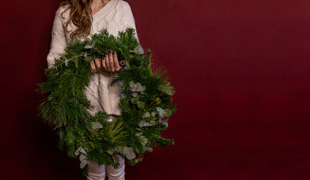Banner. Faceless young woman in knitted white sweater holds green Christmas wreath in hands on dark burgundy isolated background. Place for text. New Year Sales. Handmade interior decorations.の写真素材