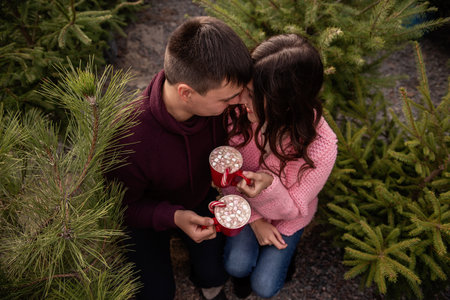 Couple in love in knitted sweaters sits among green pines, fir trees, Christmas tree market. Young man and woman drinking hot cocoa from red mugs with marshmallows, candy cane. Holiday atmosphere, funの写真素材