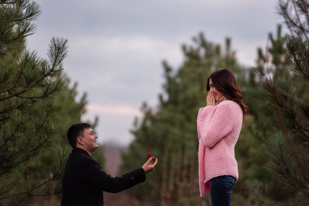 Young man in black coat kneels in front of beautiful woman, proposes marriage, holding out red box with gold ring in forest. Girl in pink knitted sweater is shocked by engagement. Authentic weddingの写真素材