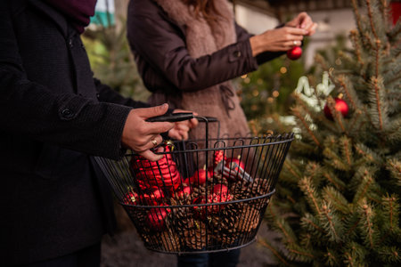 Close-up of mans hands that hold an iron basket with red balls. Defocused background the girls hands decorate the Christmas tree. Christmas atmosphere, home, backyard decoration. Faceless coupleの写真素材