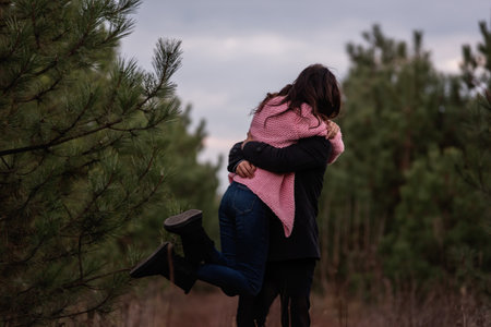 Happy couple spins in coniferous forest on path. Young man tightly hugs curly-haired woman in pink sweater. Winter engagement, autumn wedding. Proposal of marriage. Lifestyle, countryside. Copy spaceの写真素材