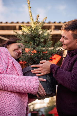 Couple in love chooses Christmas tree in outdoor market. Young man and beautiful woman look at each other, holding tub with live Christmas tree in hands. Eco-friendly choice for holiday decorations.の写真素材