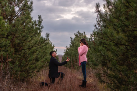 Young man in black coat kneels in front of beautiful woman, proposes marriage, holding out red box with gold ring in forest. Girl in pink knitted sweater is shocked by engagement. Authentic weddingの写真素材