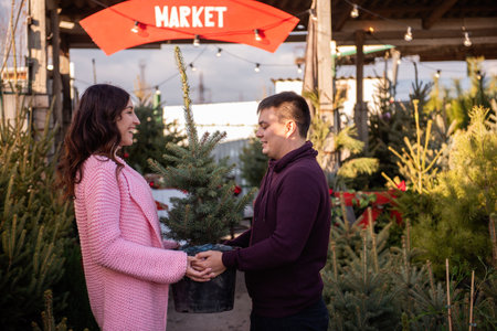 Couple in love chooses Christmas tree in outdoor market. Young man and beautiful woman look at each other, holding tub with live Christmas tree in hands. Eco-friendly choice for holiday decorations.の写真素材