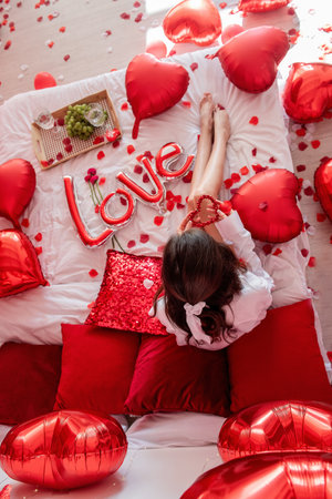 Top view of young woman sitting on bed around red balloons, rose petals. Faceless blogger holds heart-shaped beads. Decoration with inscription Love, pillows, wooden tray with two glasses and grapes.の写真素材