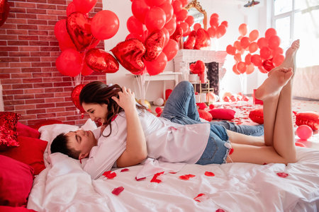 Couple in love hugs tightly on white bed among red balloons, pillows, hearts, rose petals. Man and woman look at each other, spend Valentines Day home in relaxed atmosphere. Surprise gift. Lifestyleの写真素材