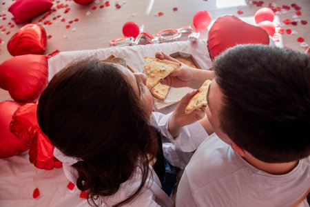 Top view of young couple feeding each other, enjoying pizza from takeaway box, with glasses of champagne, green grapes. Man and woman sharing festive, intimate moment together on a bed. Valentines dayの写真素材
