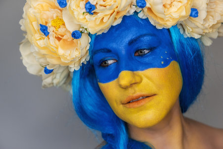 Creative close-up portrait of a young woman with yellow and blue face art, a wreath of peonies on her head on an isolated gray background. Concept for Flag Day in Ukraine. Copy spaceの写真素材