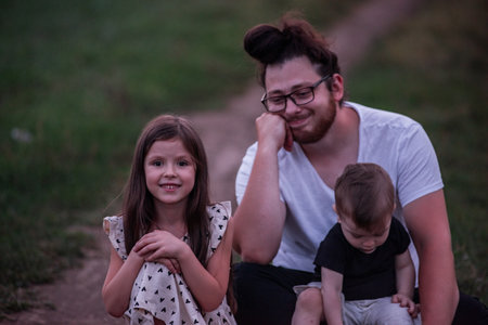 In the dimming light of dusk, father sits on rural trail, his children nestled in his arms, picture of protective love, quiet contemplation. Little girl and small boy playing near manの写真素材