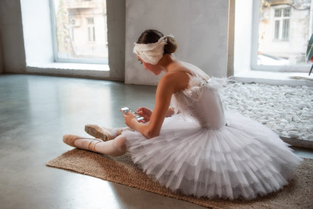 Young ballerina in white tutu, with feather tiara, sits on the floor in loft interior, holds smartphone in hands, surfing the Internet. Teenage social media. Break between rehearsals, lifestyleの写真素材