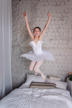 Joyful Young Ballerina Jumping on Bed. Young girl in white swan tutu leaps joyfully on bed, fooling around, smile radiating pure happiness in cozy bedroom setting.の写真素材