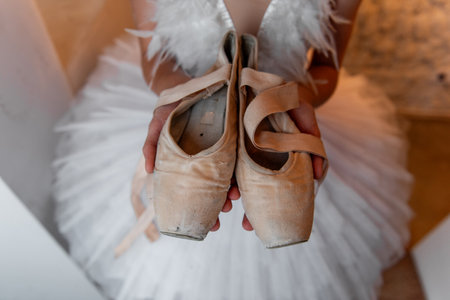 Young ballerina in white tutu, feathered hair tiara accessory stands thoughtfully, holding worn pointe shoes, evoking sense of dedication and the quiet before performance. White Swanの写真素材