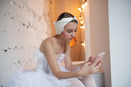 Close up portrait of young ballerina in white tutu, feather tiara, sits on the steps in loft interior, holds smartphone in hands, surfs the Internet. Teenager social media. Break between rehearsalsの写真素材