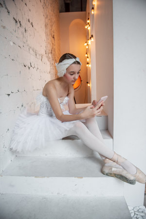 Young ballerina in white tutu, with feather tiara, sits on the steps in loft interior, holds smartphone in hands, surfs the Internet. Teenager social media. Break between rehearsals. Copy spaceの写真素材