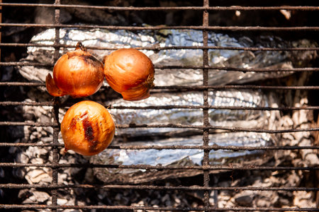 Close-up of three whole onions roasting on a grill outdoors, with foil-wrapped food beneath. Perfect for camping and outdoor cooking.の写真素材