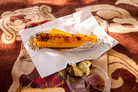 Close-up of a grilled corn on the cob wrapped in foil, served with a roasted sweet potato and onion on a patterned blanket. Perfect for outdoor picnics or barbecues.の写真素材