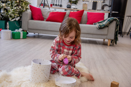 Joyful child in plaid pajamas unwraps a surprise toy from a gift box, sitting on a cozy rug in a festive living room. Christmas morningの写真素材