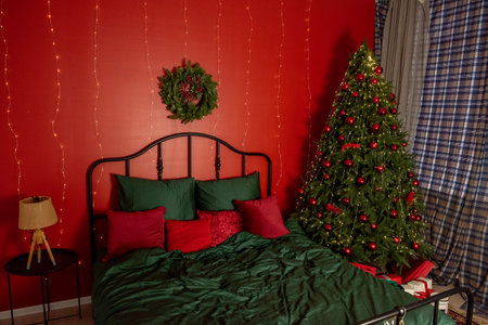 Warm inviting bedroom interior with festive Christmas decorations, lit tree adorned with ornaments. Room style with red green colors, wreath, string lights garland on walls, cozy holiday atmosphereの写真素材