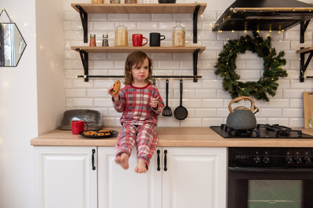 Little girl in plaid pajamas sits on kitchen Christmas counter, holding cookie. Holiday decorations, cozy atmosphere. Scandinavian modern interior, decorated with Christmas wreath, twinkling lightsの写真素材