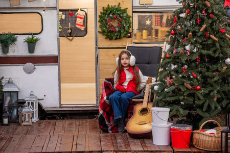 Young girl wearing earmuffs sitting outdoors beside a decorated Christmas tree, guitar, and holiday...