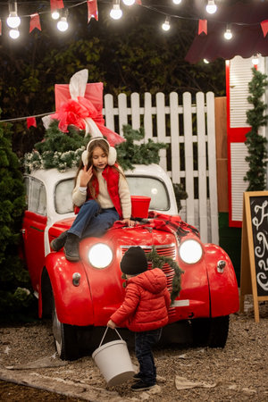 Young girl in earmuffs sits on a red car decorated for Christmas while her toddler brother stands nearby holding a bucket, both dressed warmly for a cozy holiday scene.の写真素材