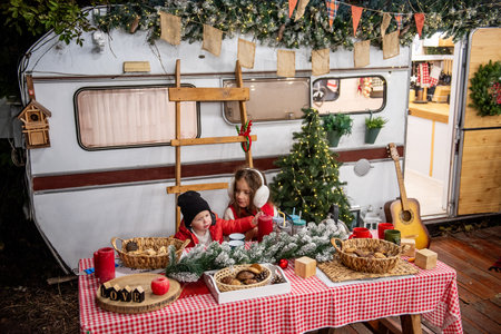 Two children dressed warmly sit at a festive outdoor table with snacks, surrounded by holiday decor, including Christmas lights and a tree, in front of a cozy caravan.の写真素材