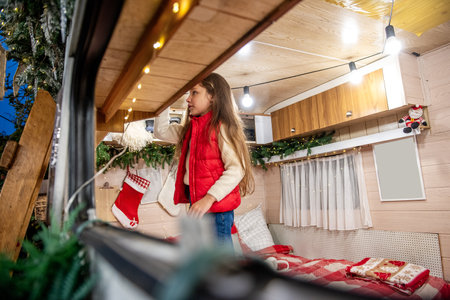 Girl in a red vest decorates a festive camper filled with Christmas stockings, lights, and greenery. She stands in the cozy interior, preparing for the holiday season.の写真素材