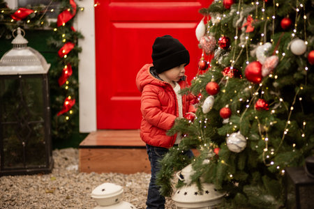 Smiling toddler boy in red jacket stands beside decorated vintage car with a wreath on the grille. A "Happy New Year" sign and festive decorations create a charming holiday scene. Back yardの写真素材