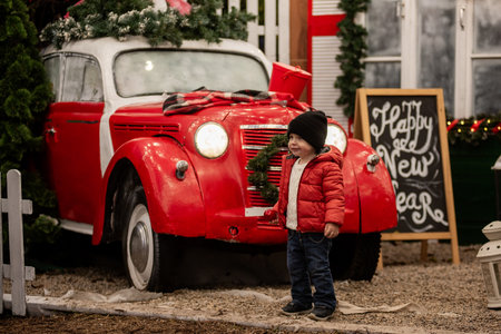 Smiling toddler boy in red jacket stands beside decorated vintage car with a wreath on the grille. A "Happy New Year" sign and festive decorations create a charming holiday scene. Back yardの写真素材