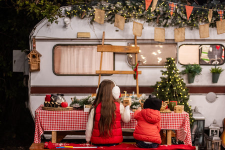 Two children in warm winter clothing sitting at a festive table outdoors, celebrating Christmas by a decorated camper van with lights and seasonal decorations.の写真素材