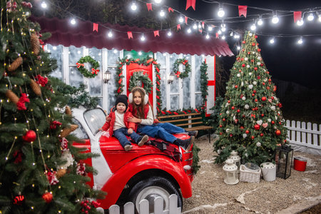 Two siblings boy and girl sit on a vintage red car in front of a festive house decorated with wreaths, garlands, and Christmas lights. The cozy winter scene exudes holiday cheer.の写真素材