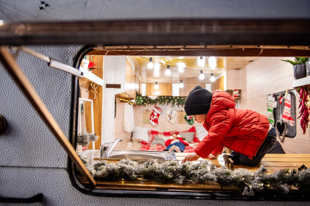 Joyful toddler boy in a red jacket sits on a wooden counter in a decorated Christmas camper, while their sibling relaxes on a bed in the background. Cozy holiday vibes in a mobile home.の写真素材