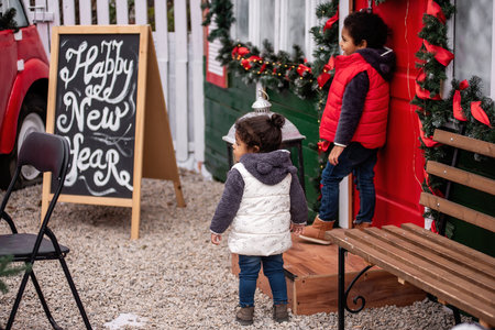 Little African American siblings explore festive decorations by a Christmas tree outdoors, surrounded by holiday ornaments, lights, and a red wreath-adorned door.の写真素材