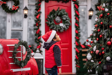 Little African American boy in a Santa hat and red vest gazes up in wonder at holiday decorations, surrounded by a beautifully adorned tree, garlands, and a vintage truck with a wreath.の写真素材