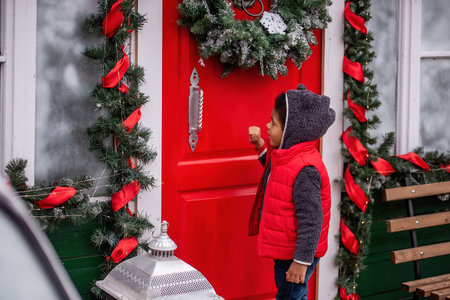 Cheerful boy in a cozy bear hoodie and red vest knocks on a festive red door adorned with holiday wreaths and garlands, capturing the magic of the Christmas season.の写真素材