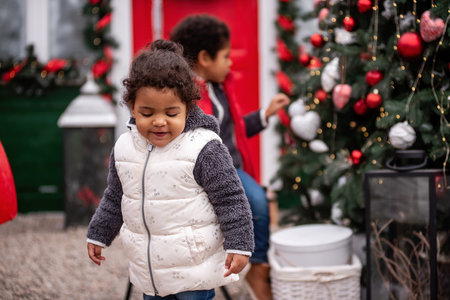 Little African American siblings explore festive decorations by a Christmas tree outdoors, surrounded by holiday ornaments, lights, and a red wreath-adorned door.の写真素材