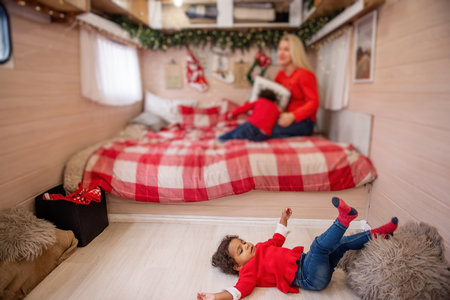 Cheerful diversity family shares a playful moment in a holiday-decorated camper. Little girl smiles brightly, while mother and brother engage in a fun game on the bed.の写真素材