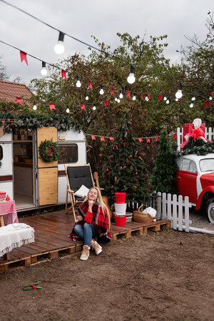 Woman in cozy plaid blanket sits outside a holiday-decorated camper. Christmas tree, guitar, and rustic holiday decor create a warm and inviting festive atmosphere.の写真素材