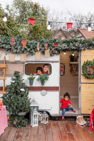Diversity Family enjoys festive time around decorated camper. Mother and son peek from window, while young girl sits at door. Holiday greenery, lights, and ornaments enhance the cozy outdoor settingの写真素材