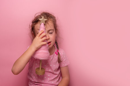 Young serious girl playfully holding a pink cotton candy-like Christmas tree with a glittering star topper against a pastel pink background. Perfect for creative and festive themes.の写真素材