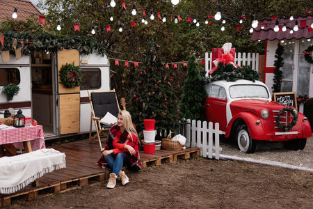 Woman in cozy plaid blanket sits outside a holiday-decorated camper. Christmas tree, guitar, and...