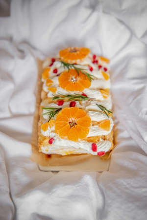 Beautifully presented dessert featuring orange slices, pomegranate seeds, cream, rosemary garnish on parchment, surrounded by soft white fabric for a cozy feel. Meringue Roll with Orange Curd dessertの写真素材